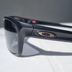 Puede incluir: Primer plano de gafas de sol negras con lentes grises oscuras. Las gafas de sol tienen un dise&ntilde;o elegante y moderno con un logotipo met&aacute;lico en la patilla. La montura es de color negro brillante y las lentes parecen polarizadas.