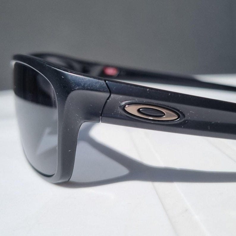 Puede incluir: Primer plano de gafas de sol negras con lentes grises oscuras. Las gafas de sol tienen un dise&ntilde;o elegante y moderno con un logotipo met&aacute;lico en la patilla. La montura es de color negro brillante y las lentes parecen polarizadas.