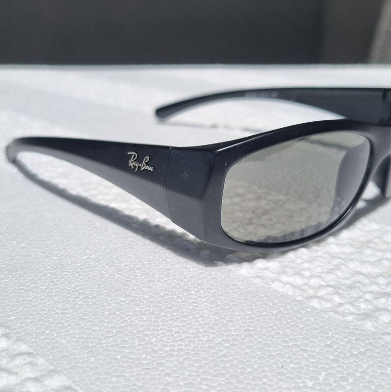 May include: Black sunglasses with a wrap-around frame design and grey tinted lenses. The temples feature the silver Ray-Ban logo on the side. These sunglasses have a smooth matte finish and a curved shape that contours to the face for coverage.