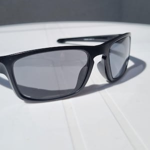 Puede incluir: Gafas de sol negras con montura rectangular y lentes grises. Las gafas de sol est&aacute;n hechas de un material negro mate y tienen un dise&ntilde;o cl&aacute;sico y deportivo. Las lentes son reflectantes y est&aacute;n sobre una superficie blanca.