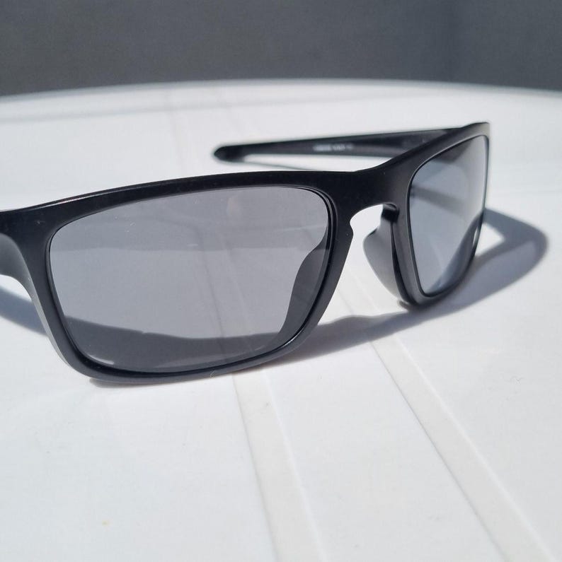 Puede incluir: Gafas de sol negras con montura rectangular y lentes grises. Las gafas de sol est&aacute;n hechas de un material negro mate y tienen un dise&ntilde;o cl&aacute;sico y deportivo. Las lentes son reflectantes y est&aacute;n sobre una superficie blanca.