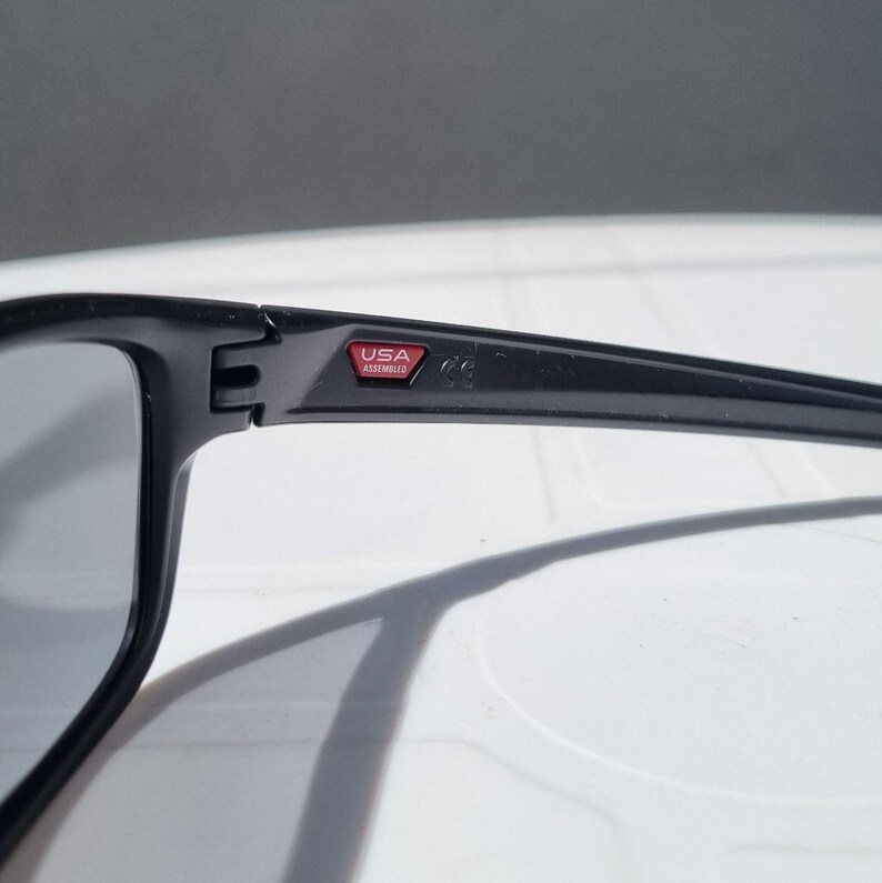 Puede incluir: Gafas de sol negras con lentes grises. La montura es gruesa y angular, con un peque&ntilde;o emblema rojo y blanco "USA Assembled" en la patilla. Las gafas de sol est&aacute;n dise&ntilde;adas para uso en exteriores y ofrecen protecci&oacute;n solar.