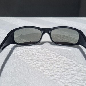 May include: A pair of black sunglasses featuring a wrap-around frame design with dark, tinted lenses. The frame has a smooth matte finish and curved temples that extend outward, providing a wide field of vision through the connected lens structure.