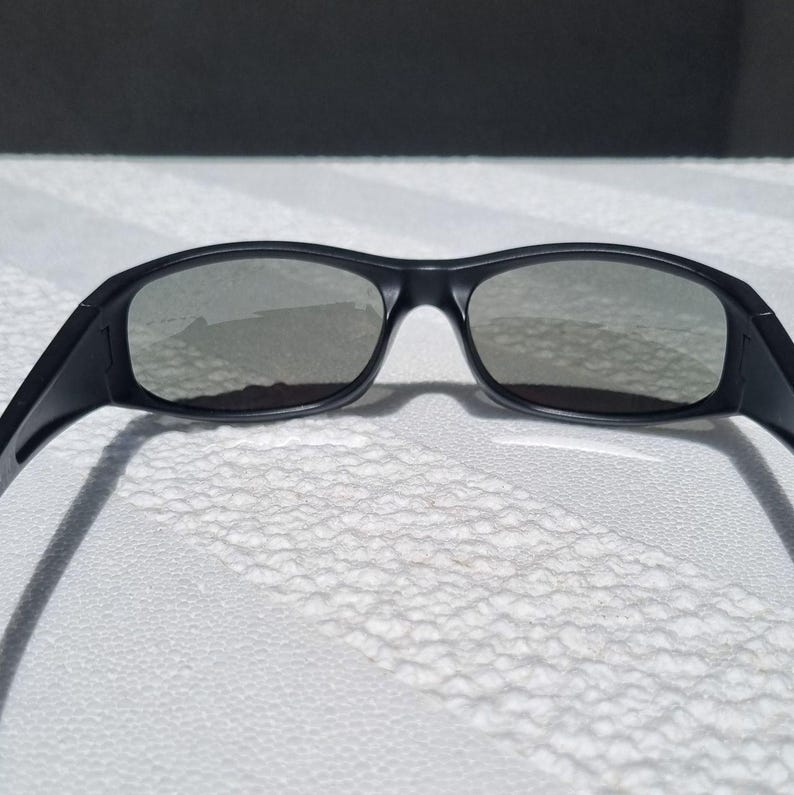 May include: A pair of black sunglasses featuring a wrap-around frame design with dark, tinted lenses. The frame has a smooth matte finish and curved temples that extend outward, providing a wide field of vision through the connected lens structure.
