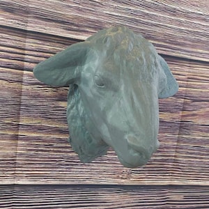 May include: A gray sheep head wall sculpture. The sculpture is mounted on a wooden wall with a natural wood grain pattern. A small potted plant with green grass sits on a wooden surface below the sculpture.