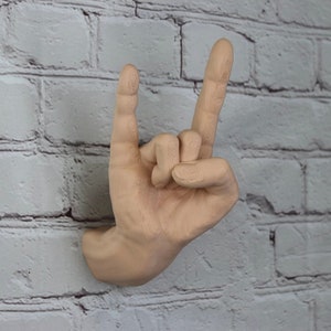 Könnte beinhalten: Eine beige Handskulptur in Form einer Rock'n'Roll-Handgeste. Die Hand ist an einer weißen Backsteinwand montiert.