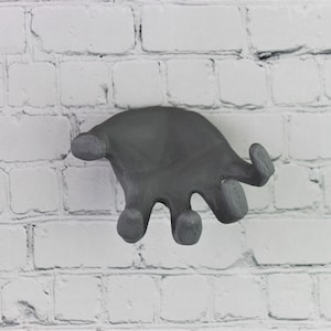 May include: A gray hand-shaped wall hook, designed to look like a hand reaching out, mounted on a white brick wall.