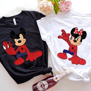 May include: Two t-shirts, one black and one white, each featuring a cartoon character in a Spider-Man costume. The black shirt has Mickey Mouse, and the white shirt has Minnie Mouse. Both characters are in a dynamic pose with red and blue accents.