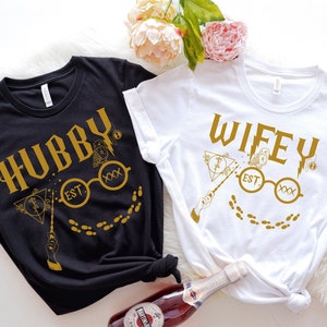 以下が含まれることがあります： 金色のグラフィックが施された黒と白のTシャツ。黒いシャツには「Hubby」、白いシャツには「Wifey」と書かれています。どちらのシャツにも、杖、メガネ、足跡のデザインがあり、「EST」とローマ数字が描かれています。