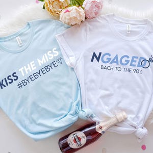 Op de afbeelding: Twee T-shirts, één lichtblauw en één wit, met tekst voor een vrijgezellenfeest. Het lichtblauwe shirt zegt "KISS THE MISS #BYEBYEBYE" en het witte shirt zegt "NGAGED Bach to the 90's". Een fles champagne staat in de afbeelding.