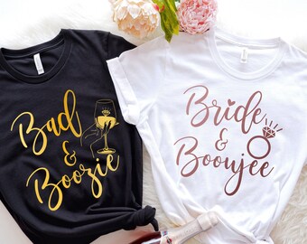 May include: Two white t-shirts with black text. The shirt on the left says "Bad & Boozie" in gold glitter lettering with a gold glitter glass of wine. The shirt on the right says "Bride & Boozy" in brown glitter lettering with a brown glitter diamond ring.