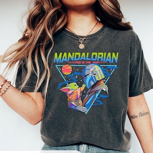 May include: A dark grey t-shirt with a retro-style graphic of the Mandalorian and Grogu. The graphic features a triangle with a planet and stars, and the text "Mandalorian - This is the Way".