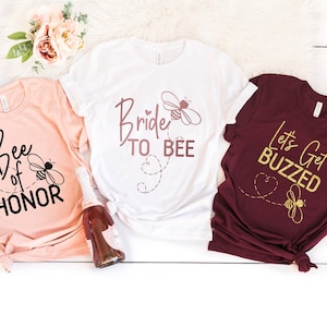 May include: Three t-shirts with bee-themed designs for a bridal party. The white t-shirt says "Bride to Bee" in pink script with a bee graphic. The peach t-shirt says "Bee of Honor" in black with a bee graphic. The maroon t-shirt says "Let's Get Buzzed" in gold with a bee graphic.