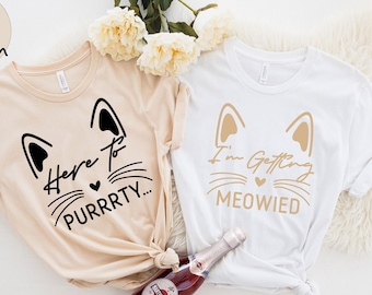 May include: Two t-shirts with cat-themed designs for a bridal party. One shirt is beige with black text that says "Here to purrrty..." and the other shirt is white with gold text that says "I'm getting meowied."