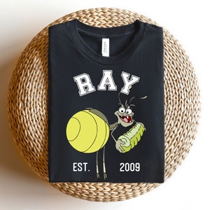 Puede incluir: Camiseta negra con una imagen de dibujos animados de Ray de La Princesa y el Sapo de Disney. El texto "RAY" est&aacute; en blanco con un contorno blanco. El texto "EST. 2009" tambi&eacute;n est&aacute; en blanco.