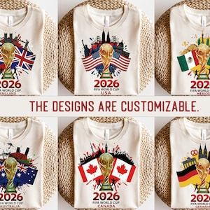 May include: Cream t-shirts showcasing designs for the 2026 FIFA World Cup. Each shirt features a golden trophy, flags, and cityscapes representing England, USA, Mexico, Australia, Canada, and Germany. The text "2026 FIFA WORLD CUP" is displayed.