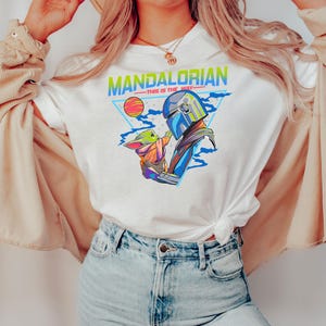 May include: White t-shirt with a retro-style graphic of the Mandalorian and Grogu. The graphic features the text "Mandalorian - This is the Way" in a colorful, pixelated font.