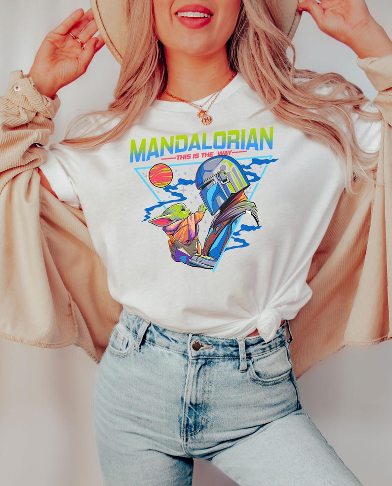 May include: White t-shirt with a retro-style graphic of the Mandalorian and Grogu. The graphic features the text "Mandalorian - This is the Way" in a colorful, pixelated font.