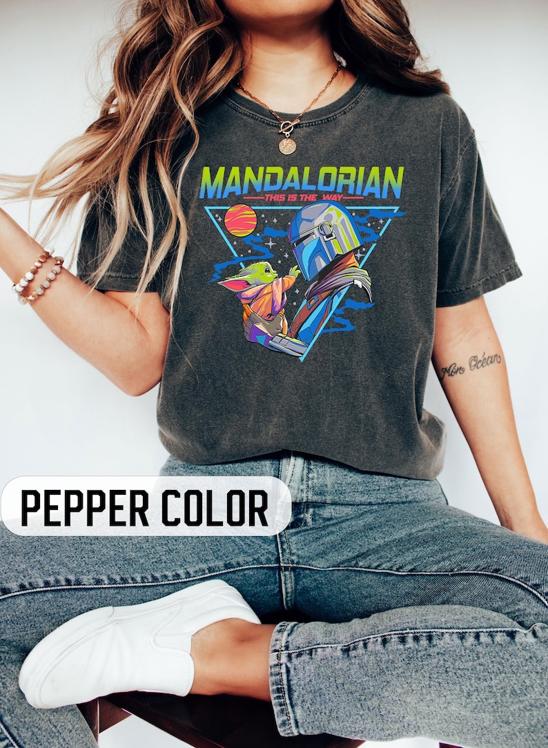 May include: A dark grey t-shirt with a retro-style graphic of the Mandalorian and Grogu. The graphic features a triangle with a planet and stars, and the text "Mandalorian - This is the Way".
