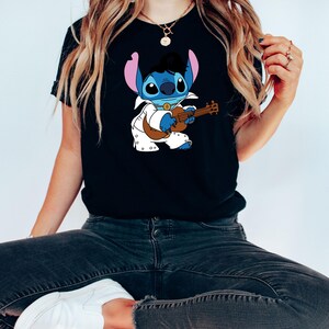 May include: Black t-shirt featuring a blue cartoon character playing a guitar. The character is wearing a white jumpsuit with a black belt and a black pompadour hairstyle.