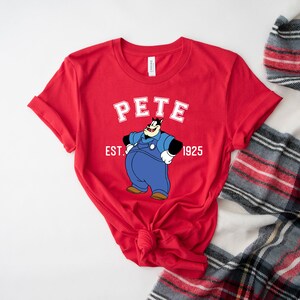 Puede incluir: Camiseta roja de manga corta con una imagen de dibujos animados de Pete el cachorro con un mono y el texto "Pete Est. 1925".
