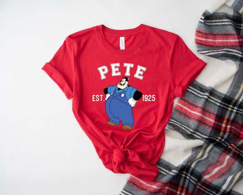 Puede incluir: Camiseta roja de manga corta con una imagen de dibujos animados de Pete el cachorro con un mono y el texto "Pete Est. 1925".