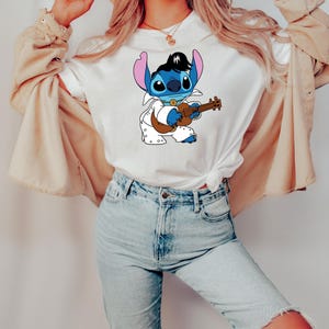 May include: A white t-shirt with a blue cartoon character playing a guitar. The character is wearing a white jumpsuit with a black belt and has a black pompadour hairstyle. The character is surrounded by white polka dots.