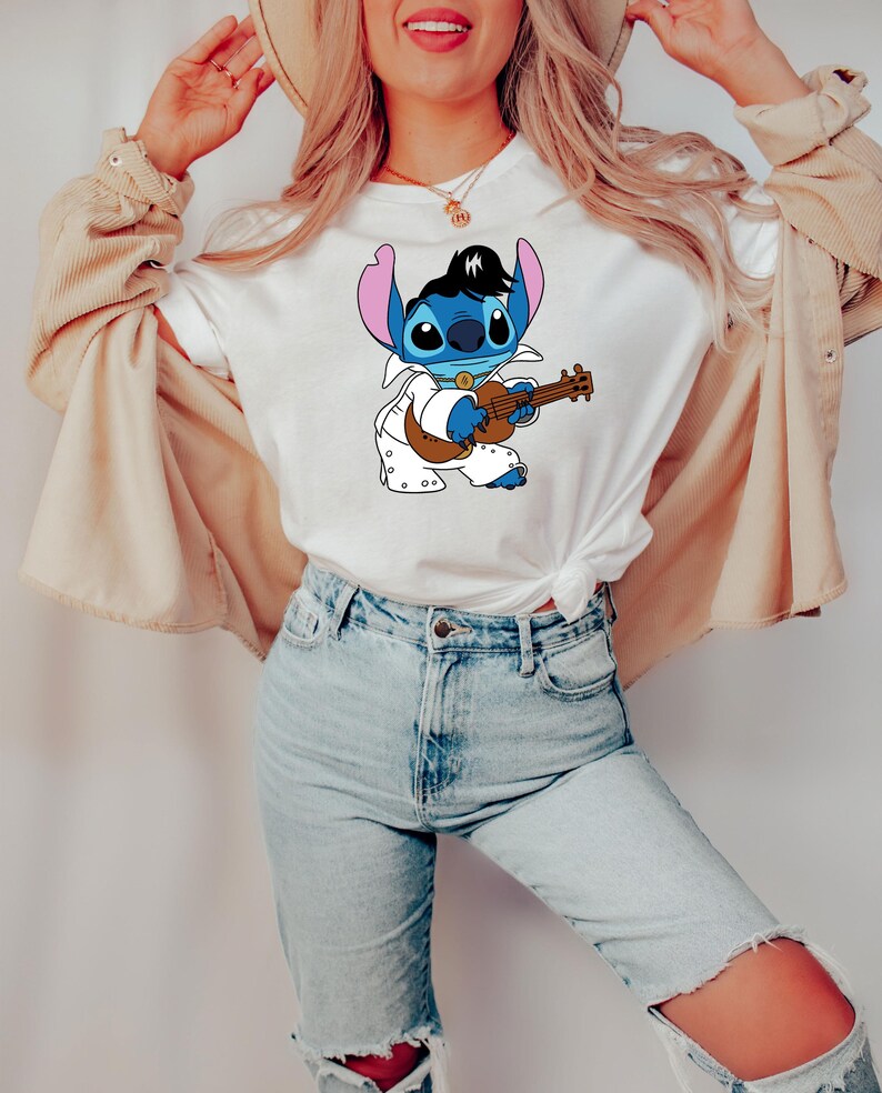 May include: A white t-shirt with a blue cartoon character playing a guitar. The character is wearing a white jumpsuit with a black belt and has a black pompadour hairstyle. The character is surrounded by white polka dots.