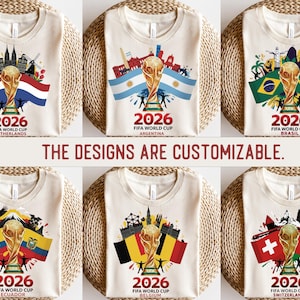 May include: A collection of cream-coloured t-shirts, each showcasing a FIFA World Cup 2026 design. The shirts display a golden trophy, flags, and cityscapes representing the Netherlands, Argentina, Brazil, Ecuador, Belgium, and Switzerland. Text: "THE DESIGNS ARE CUSTOMIZABLE."