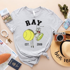 Puede incluir: Camiseta gris con una imagen de dibujos animados de Ray de 'Up' de Disney sosteniendo un pepinillo. El texto 'RAY EST. 2009' est&aacute; impreso en la camiseta.