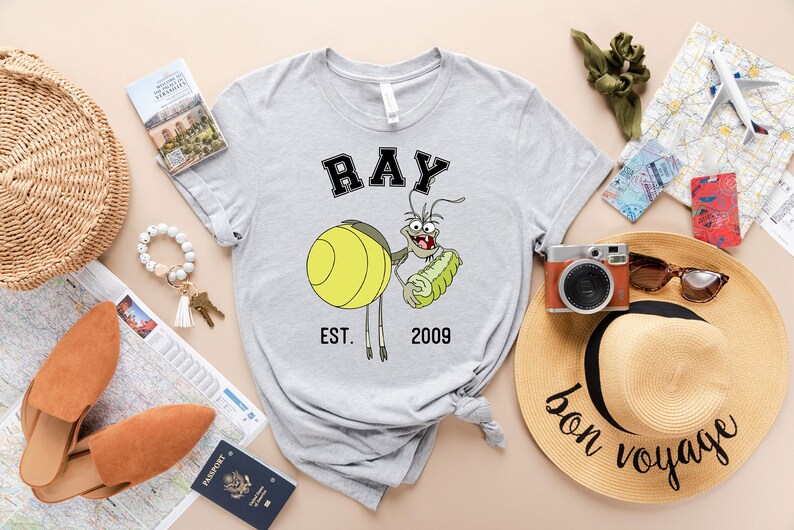 Puede incluir: Camiseta gris con una imagen de dibujos animados de Ray de 'Up' de Disney sosteniendo un pepinillo. El texto 'RAY EST. 2009' est&aacute; impreso en la camiseta.