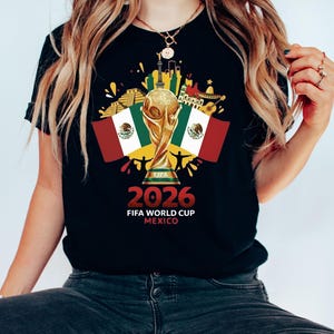 May include: Black t-shirt with a gold FIFA World Cup trophy, Mexican flags, and the text "2026 FIFA WORLD CUP MEXICO". The design incorporates Mexican cultural symbols and a cityscape.