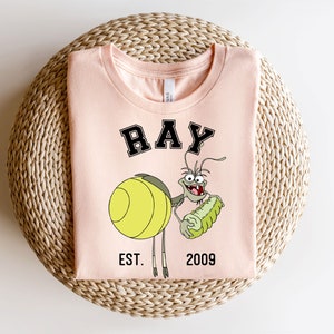 Puede incluir: Una camiseta rosa con un personaje de dibujos animados de Ray de La Sirenita de Disney. El personaje sostiene un pepinillo verde y tiene el texto "RAY EST. 2009" impreso en la camiseta.