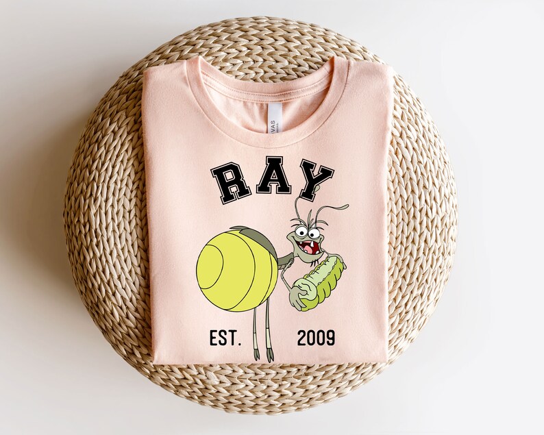 Puede incluir: Una camiseta rosa con un personaje de dibujos animados de Ray de La Sirenita de Disney. El personaje sostiene un pepinillo verde y tiene el texto "RAY EST. 2009" impreso en la camiseta.