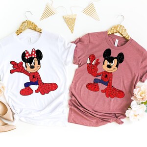 May include: Two t-shirts, one white and one mauve, each featuring a cartoon character dressed as Spider-Man. The white shirt has Minnie Mouse, and the mauve shirt has Mickey Mouse. Both characters are in a Spider-Man pose, with red and blue outfits.