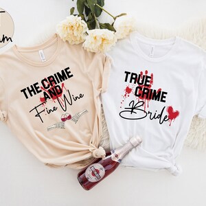 Pu&ograve; includere: Due magliette bianche con testo nero e schizzi di sangue rosso. Il testo sulla maglietta beige dice "The Crime and Fine Wine" con una mano scheletrica che tiene un bicchiere di vino. Il testo sulla maglietta bianca dice "True Crime Bride".