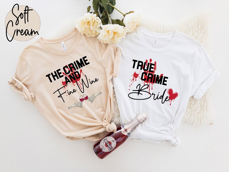 Pu&ograve; includere: Due magliette bianche con testo nero e schizzi di sangue rosso. Il testo sulla maglietta beige dice "The Crime and Fine Wine" con una mano scheletrica che tiene un bicchiere di vino. Il testo sulla maglietta bianca dice "True Crime Bride".