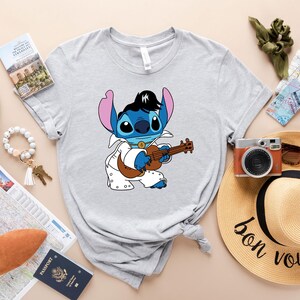 May include: A gray t-shirt with a blue cartoon character playing a guitar. The character is wearing a white jumpsuit and has a black pompadour hairstyle. The text "bon voyage" is written on a straw hat in the background.