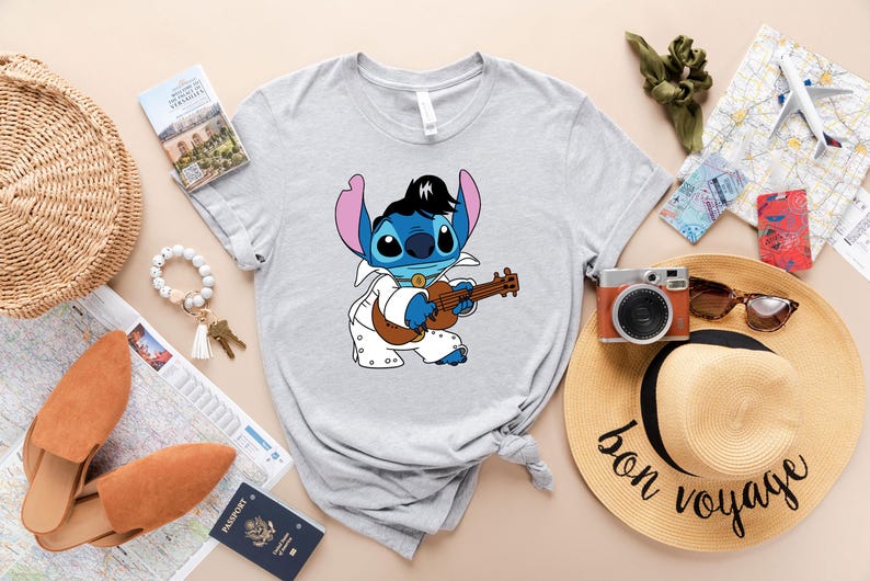 May include: A gray t-shirt with a blue cartoon character playing a guitar. The character is wearing a white jumpsuit and has a black pompadour hairstyle. The text "bon voyage" is written on a straw hat in the background.