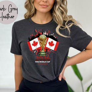 May include: Dark grey heather t-shirt with a graphic celebrating the 2026 FIFA World Cup in Canada. The design includes the World Cup trophy, Canadian flags, and the text "2026 FIFA WORLD CUP CANADA".