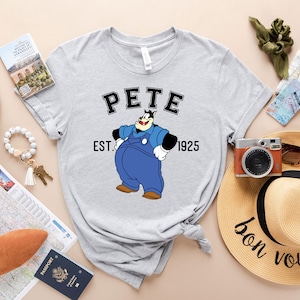 Puede incluir: Una camiseta gris con una imagen de dibujos animados de Pete el perro de las caricaturas de Mickey Mouse. La camiseta tiene el texto "Pete" en una fuente universitaria en blanco y negro con el texto "Est. 1925" debajo de la imagen.
