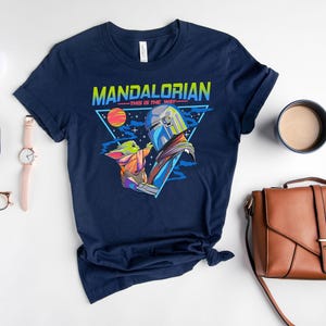 May include: A navy blue t-shirt with a retro-style graphic of the Mandalorian and Grogu. The graphic features a triangle with the words "Mandalorian - This is the Way -" and a colorful image of the Mandalorian holding Grogu.