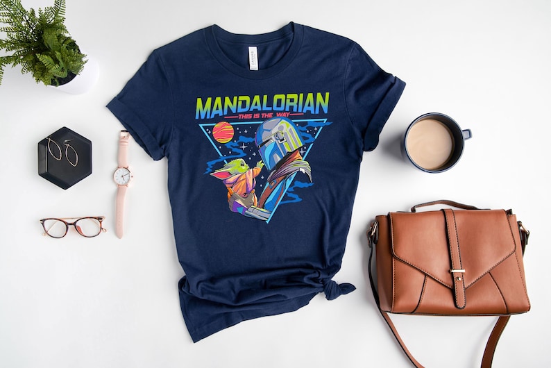 May include: A navy blue t-shirt with a retro-style graphic of the Mandalorian and Grogu. The graphic features a triangle with the words "Mandalorian - This is the Way -" and a colorful image of the Mandalorian holding Grogu.