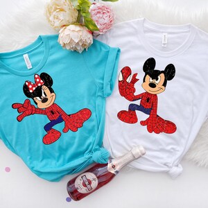 May include: Two t-shirts, one turquoise and one white, each with a cartoon character in a Spider-Man-inspired costume. The turquoise shirt features Minnie Mouse, and the white shirt features Mickey Mouse. A bottle of sparkling wine is in the foreground.