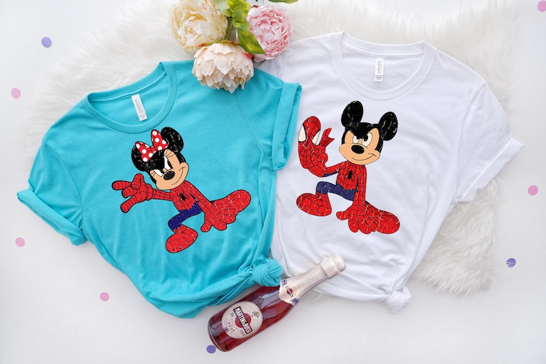 May include: Two t-shirts, one turquoise and one white, each with a cartoon character in a Spider-Man-inspired costume. The turquoise shirt features Minnie Mouse, and the white shirt features Mickey Mouse. A bottle of sparkling wine is in the foreground.