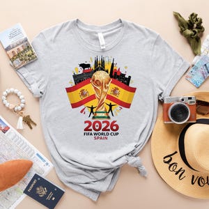 May include: A light grey t-shirt with a graphic for the 2026 FIFA World Cup in Spain. The design features the World Cup trophy, Spanish flags, and the text "2026 FIFA WORLD CUP SPAIN". Travel accessories are also visible in the image.