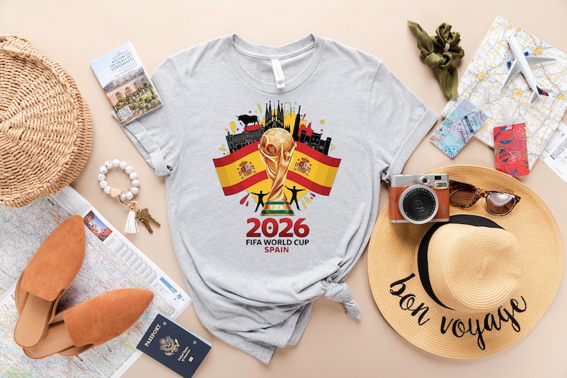 May include: A light grey t-shirt with a graphic for the 2026 FIFA World Cup in Spain. The design features the World Cup trophy, Spanish flags, and the text "2026 FIFA WORLD CUP SPAIN". Travel accessories are also visible in the image.