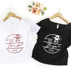 May include: Two V-neck t-shirts, one white and one black, hang on gold sequined hangers. The white shirt says "Bride" and the black shirt says "Babe", both with "Las Vegas 2023" and playing card graphics.
