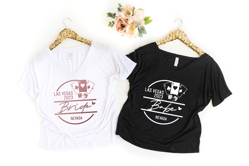 May include: Two V-neck t-shirts, one white and one black, hang on gold sequined hangers. The white shirt says "Bride" and the black shirt says "Babe", both with "Las Vegas 2023" and playing card graphics.