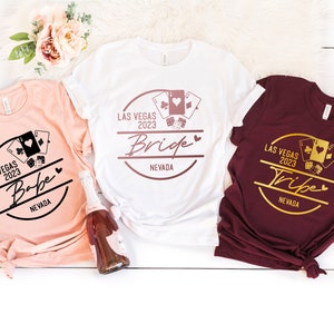 May include: Three t-shirts in heather peach, white, and maroon, each with a Las Vegas 2023 graphic. The heather peach shirt says "Babe", the white shirt says "Bride", and the maroon shirt says "Tribe".
